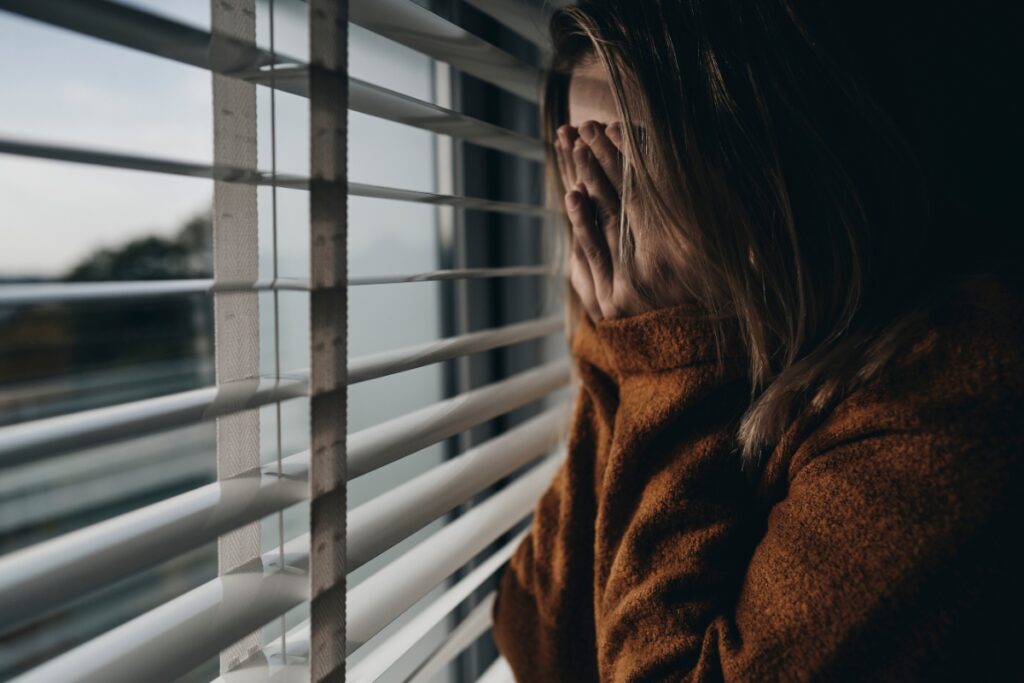 Sad and lonely woman standing at window with her hands over her face
