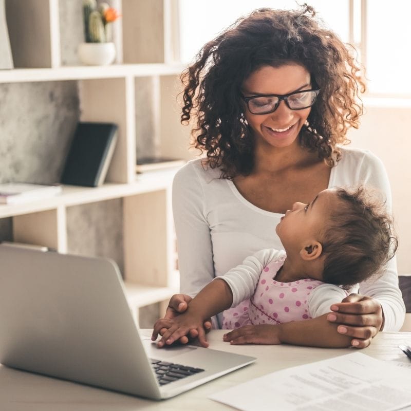 Mother with her baby on her lap as she works on her laptop. In many cases, being a stay at home mom is more financially stable than sending your baby to daycare.