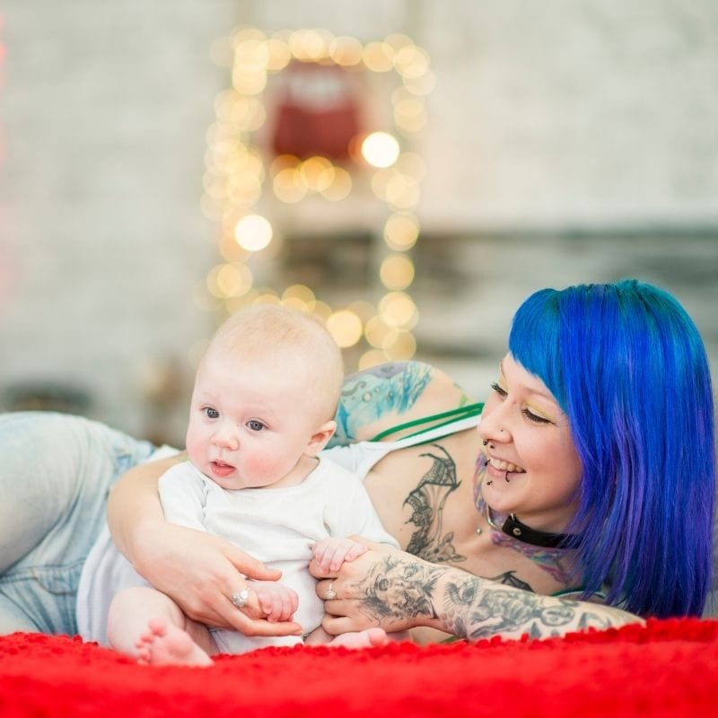 Mother with tattoos and blue hair with her cute baby in a white onesie.