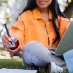 Woman in orange jacket writing in a journal with an ipad or tablet