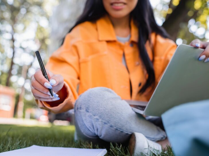 Woman in orange jacket writing in a journal with an ipad or tablet